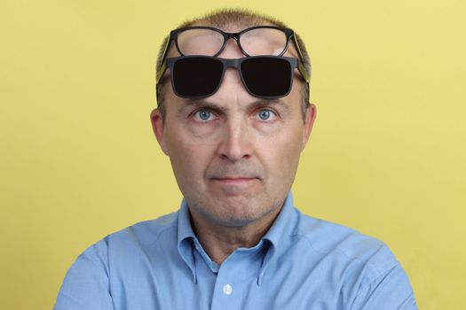 A man in a blue shirt on a yellow background with two glasses on his face.