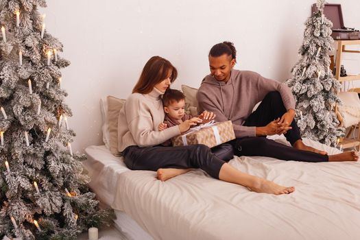 A happy multi-racial family is sitting on a big bed, in a Christmas interior, untying a ribbon on a brown gift box