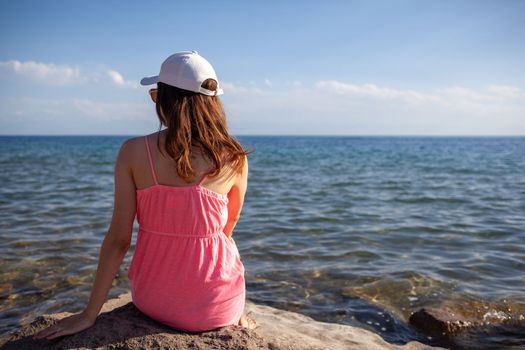 A beautiful girl in a swimsuit with long hair and a white cap looks at the blue sea. Rear view.