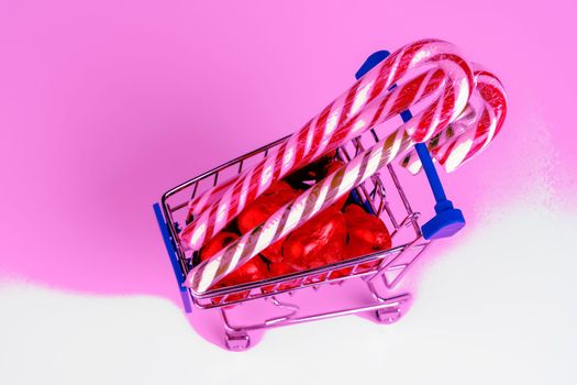 A supermarket trolley loaded with heart-shaped candies and Christmas caramel canes on a white background. Gifts and purchases for the New Year and Christmas. Online trading. A toy shopping cart carries a Christmas candy cane