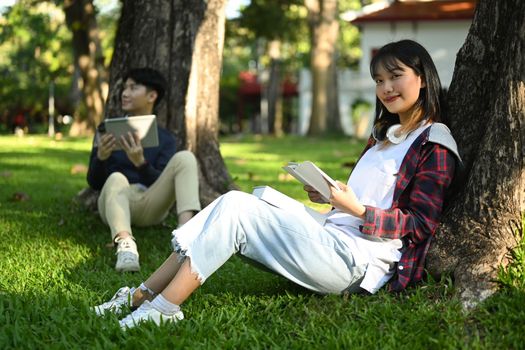 Image of smiling female student reading books with her friends at sunny beautiful garden background.