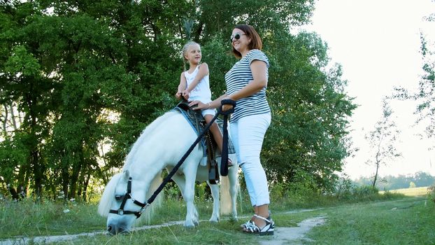 Mom and daughter are walking around the field, daughter is riding a pony, mother is holding a pony for a bridle. Cheerful, happy family vacation. Outdoors, in summer, near the forest. High quality photo