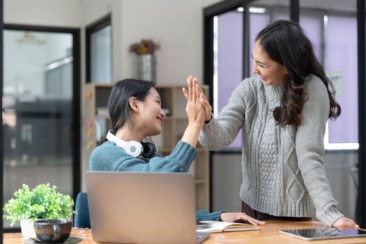 Two young Asian women show joyful expression of success at work smiling happily with a laptop computer in a modern office...