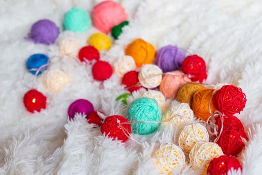A lot of colorful knitting yarn and a luminous garland in the form of balls lie on a white fluffy blanket. Cozy still life made of yarn. Close up. Copy space