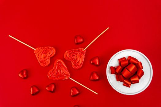 creative minimalist composition. a gift box with a ribbon and a bow, heart-shaped chocolates and heart-shaped caramel lollipops on a red background. A declaration of love. a gift and gratitude. Valentine's Day. postcard. copy space