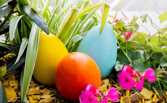 Three large multicolored wooden eggs on a flower bed, among flowers and grass. Close up. Side view