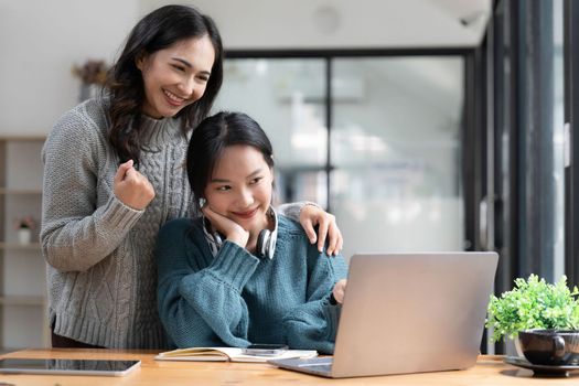 Two young Asian women show joyful expression of success at work smiling happily with a laptop computer in a modern office...