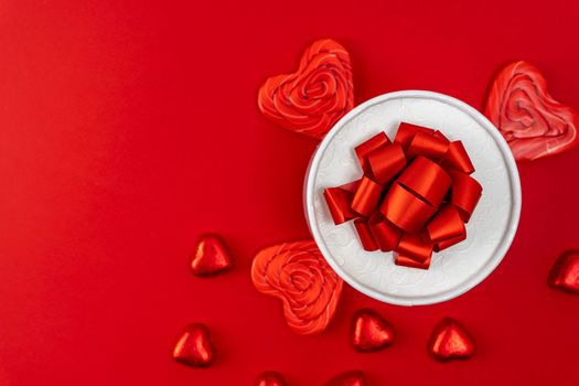 creative minimalist composition. a gift box with a ribbon and a bow, heart-shaped chocolates and heart-shaped caramel lollipops on a red background. A declaration of love. a gift and gratitude. Valentine's Day. postcard. copy space