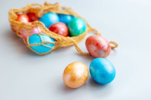 Several multi colored eggs lie in a knitted bag on white table . Three eggs lie nearby on a white background. Copy space