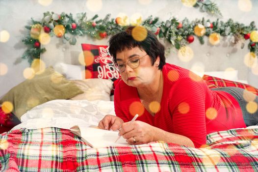 happy elderly woman with pen and notebook making wish list or to do list for new year in bed over christmas tree. elderly woman dreaming in christmas time. xmas holidays concept