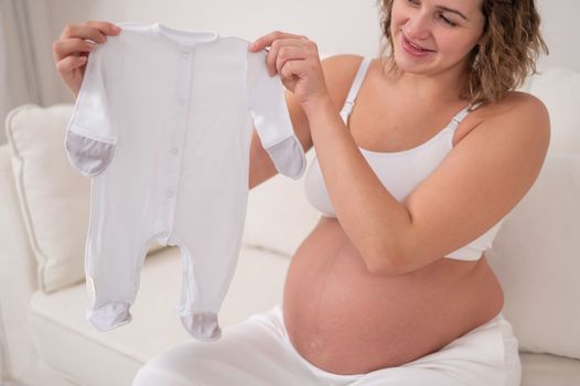 A pregnant woman sits on a white sofa and holds clothes for the expected baby