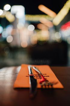 Vertical close up table setting in a restaurant. Fork and a Knife next to a napkin ready for dinner with city night lights in background.