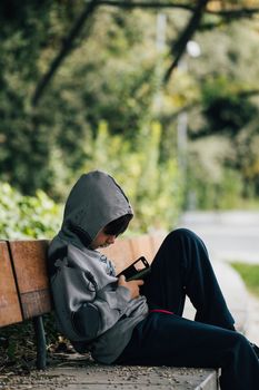 Boy with a hood sitting on a bench alone in a park looking the smartphone.