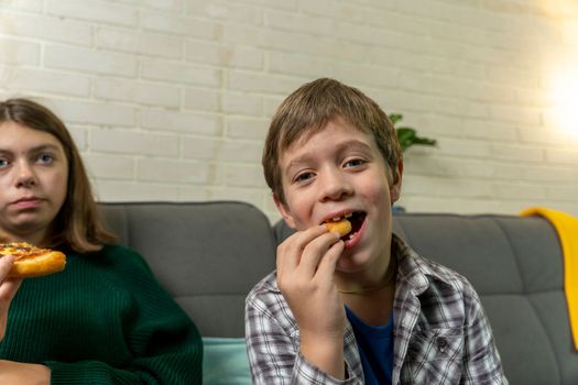 Cute Caucasian preschooler boy is sitting at home on the couch, eating pizza and watching TV. a child alone at home watching cartoons without control