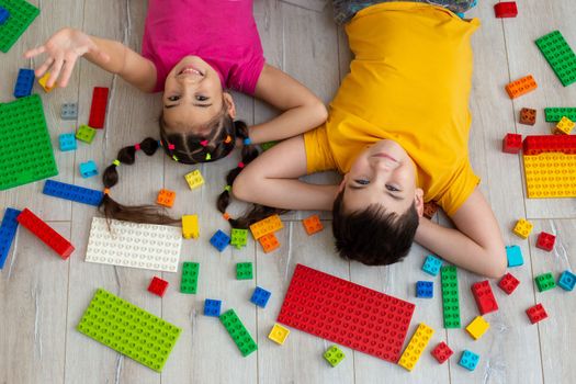 Portrait of happy boy and a little girl in bright multicolored clothes, lie on the floor, on their backs, in the room, with a multicolored plastic construction set