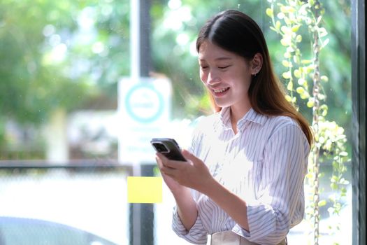 Portrait of elegant successful asian business woman speaking by smartphone and smiling happily while standing against window in office