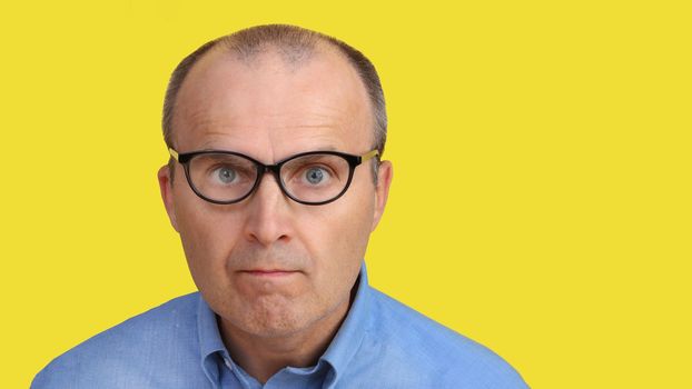Close-up of a man's face with glasses. A man with a funny look in glasses and a blue shirt on a yellow background. The man with the glasses looks at the camera.