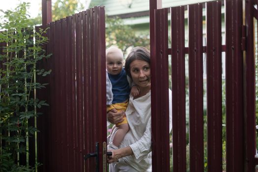 Curious woman with a baby peeps openint the gate of fence, she looks with interest wwhat her neighbor is doing