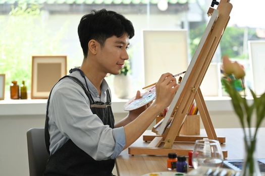 Concentred man painting picture on canvas with oil paints in bright art studio. Leisure activity and art concept.