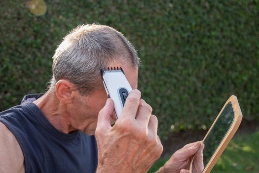 a gray-haired middle-aged man shaves his hair with a clipper in a garden on a green lawn and looks into a table mirror. High quality photo