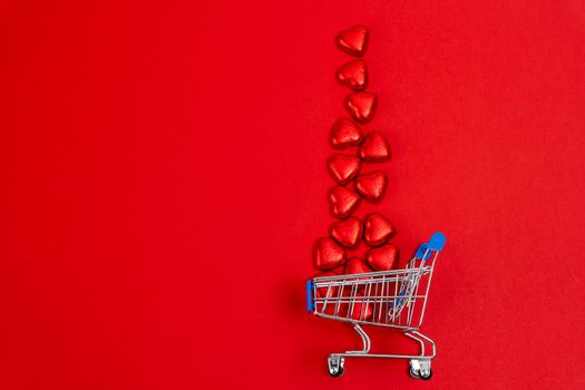 a red festive Christmas background with a supermarket cart into which heart-shaped candies wrapped in shiny foil are poured. Copy space. Red Chocolate Candy in Shape of Hearts in Mini Shopping Cart