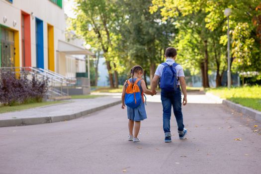 Cheerful schoolchildren, a little girl and a boy, brother and sister with backpacks hold hands go to school with multi-colored windows and green trees in the morning on a sunny summer day