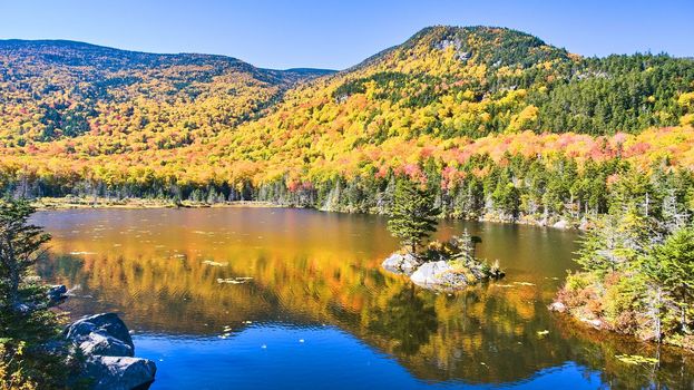 Image of Lake in New Hampshire reflecting colorful peak fall mountains and lone rocky island with pine tree