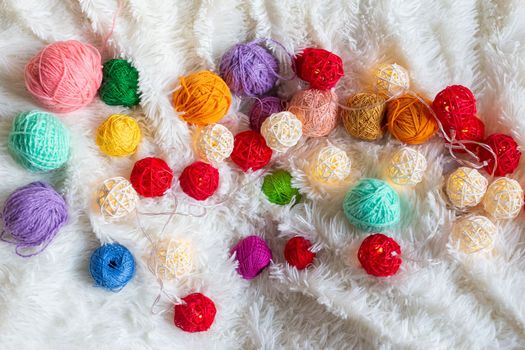 A lot of colorful knitting yarn and a luminous garland in the form of balls lie on a white fluffy blanket. Top view. Copy space