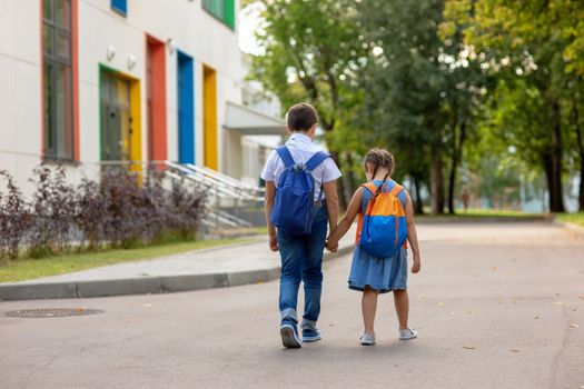 two schoolchildren, brother and sister with backpacks hold hands go to school in the morning on a sunny summer day on background school bulding . Mock up. Copy space