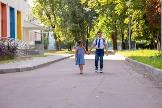 Cheerful schoolchildren, a little girl and a boy, brother and sister with backpacks hold hands go from school with multi-colored windows and green trees in the morning on a sunny summer day