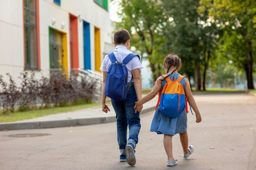 two schoolchildren, a little girl and a boy, brother and sister with backpacks hold hands go to school in the morning on a sunny summer day. Close up. Mock up