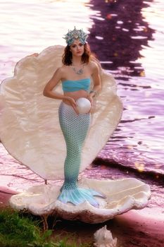 A beautiful mermaid girl in a beautiful blue diadem stands in a large white shell near the river. Holds a large round pearl