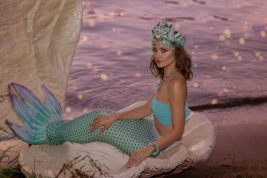 A beautiful mermaid girl in a beautiful diadem sits by the water in a large white shell. Copy space