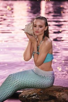 A mermaid girl sits near the water at dusk, holding a large seashell.