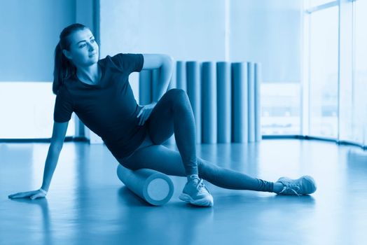 Attractive female doing foam roller exercise and posing in modern bright fitness center. Toned image.