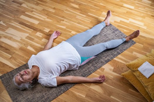 Senior caucasian woman lying on yoga mat after workout. Fit female relaxing on floor at home. 