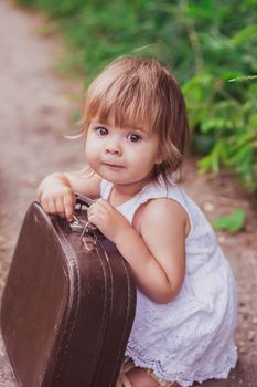 Charming baby in a white sundress tries to open an old suitcase.