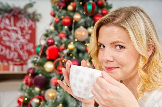 Portrait of woman 40s in white sweater holding a cup of warm coffee or tea on background of a Christmas tree. Cozy christmas atmosphere. warming non-alcoholic drink for Christmas. lifestyle