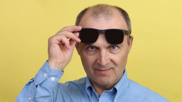 Close-up portrait of a handsome cheerful man in a blue shirt holding black glasses in his hand looking at the camera. A man with glasses on a yellow background.