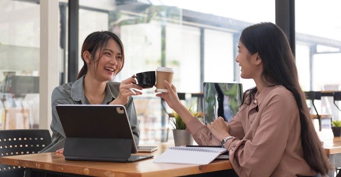 Two young Asian businesswomen show joyful expression of success at work smiling happily with a laptop computer in a modern office...