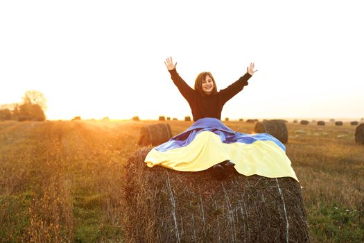 Girl with flag of Ukraine sits the field at sunset. Portrait of little girl holding a yellow and blue flag of Ukraine on a background of sunset sky. Stop war in Ukraine. Russia stop war.