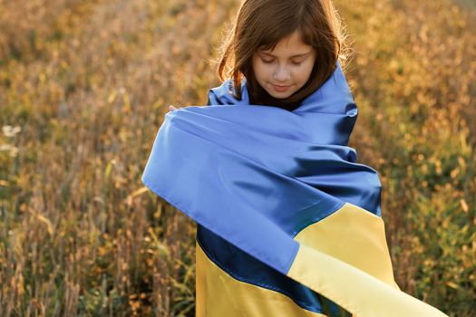 Portrait of Sad Ukrainian girl stands with a flag of Ukraine in the middle of field against a sunset sky. 8 years old girl holding the flag of Ukraine. War in Ukraine. Support for Ukraine