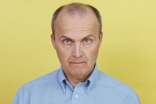 A handsome man with an intelligent and intent look. An elderly man in a blue shirt on a yellow background looks at the camera.