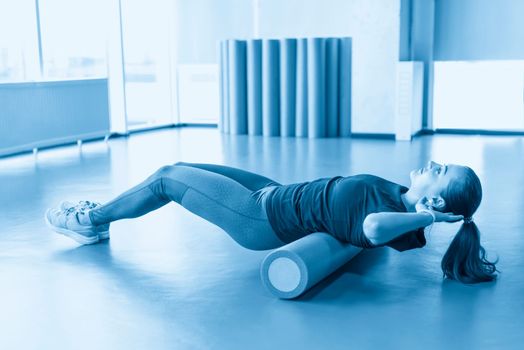 Attractive female doing foam roller exercise and posing in modern bright fitness center. Toned image.