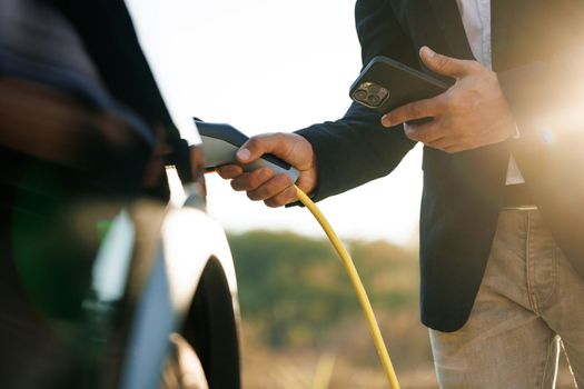 Male charging EV vehicle outdoors at sun light. Businessman unplugging zero emission electric car at charging station. Man disconnects charging cable to to electric vehicle using app on smartphone.