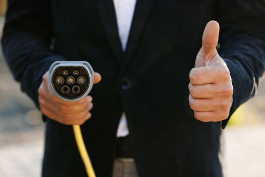 Businessman hand showing thumbs up holding power type 2 ccs cable supply plugged at electric car charging station. Man standing near electric charging station and showing thumb up.