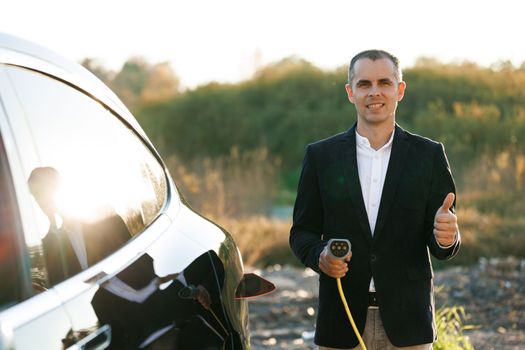 Portrait of caucasian businessman standing near electric charging station looking at camera and showing thumb up. Male holds a charging cable type 2 mennekes in his hands. Eco-friendly transport.