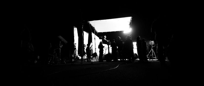 Silhouette images of video production behind the scene or b-roll or making of film shooting and movie crew team working in back of camera and lighting soft box equipment set at outdoor in the night