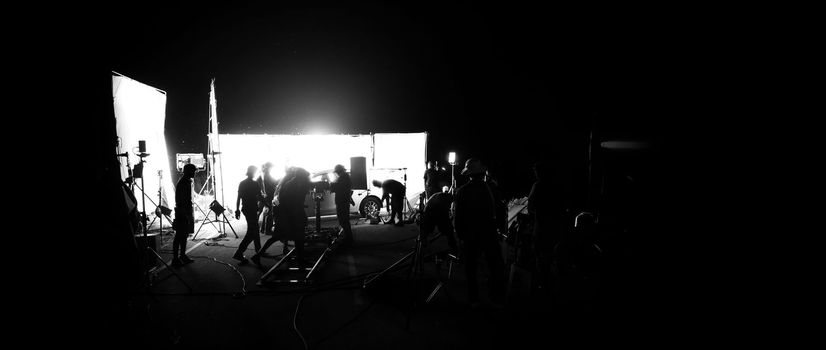 Silhouette images of video production behind the scene or b-roll or making of film shooting and movie crew team working in back of camera and lighting soft box equipment set at outdoor in the night