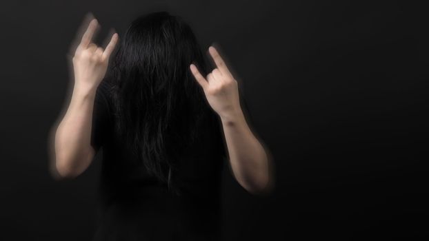 Rocker man long hair and making hand sign of rock and roll and shaking head with black color t-shirt and dark background paper in studio which represent style of music such as punk or heavy metal